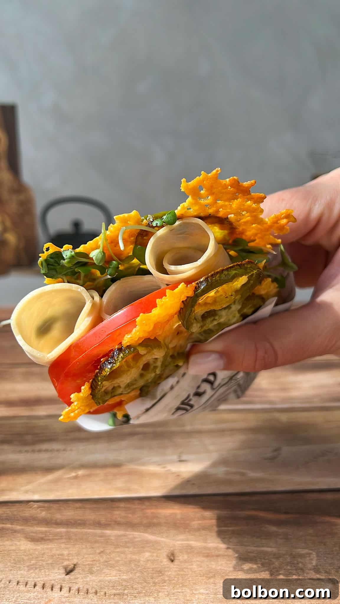 Jalapeño Hype Sandwich 2 A hand holding a sandwich with crispy cheese, pickles, tomato, greens, and rolled slices of deli meat, wrapped partially in paper, over a wooden table and gray background.