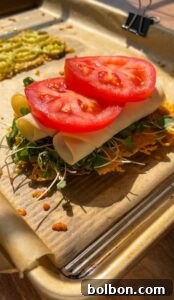 Jalapeño Hype Sandwich 7 An open-faced sandwich on parchment paper, featuring microgreens, rolled slices of cheese, and two fresh tomato slices on top of a crispy, thin base. Sunlight shines on the tray, highlighting the fresh ingredients.