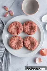 A white plate showcasing four raw meat patties, each with a visible indentation in the center, arranged on a smooth marble surface. Accompanying elements include garlic cloves, an empty bowl, a striped towel, a small dish, and cracked eggshells, illustrating the preparation for cooking.