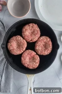 Four raw hamburger patties are arranged in a sleek black skillet, ready for cooking on a pristine marble surface. A striped cloth, garlic cloves, and empty bowls are visible nearby, highlighting the organized kitchen setting.