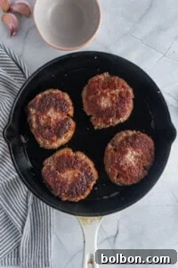 Four perfectly browned hamburger patties sizzle and cook in a black skillet, resting on a clean marble countertop. A striped cloth and a small empty bowl are positioned nearby, adding to the culinary scene.