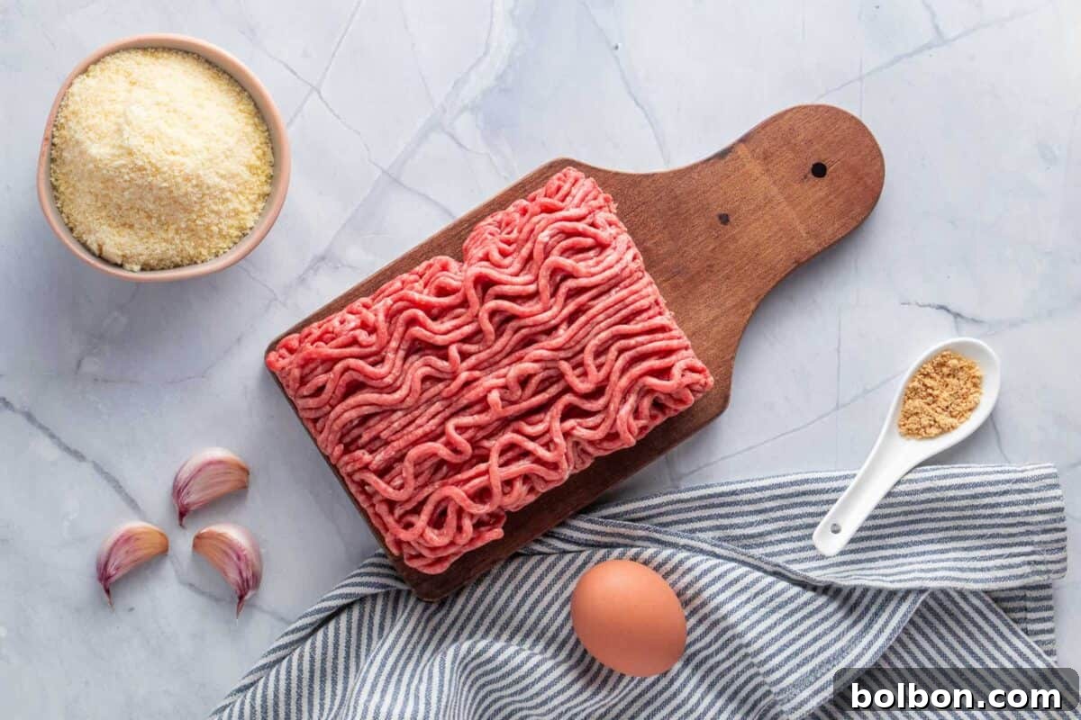 Raw ground beef on a rustic wooden cutting board, a bowl of rich grated Parmesan cheese, three fresh garlic cloves, one whole egg, a spoonful of golden breadcrumbs (though not in the final recipe mix, visually representing texture), and a striped towel on a cool marble surface.