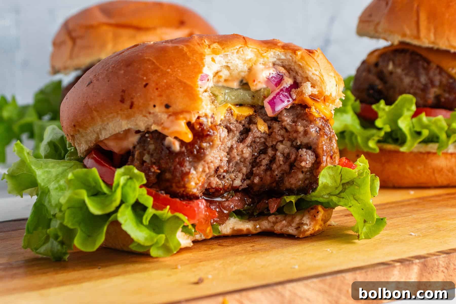 Close-up of a perfectly cooked, juicy homemade cheeseburger with a bite taken out, revealing its succulent interior. It rests on a rustic wooden board, generously topped with fresh lettuce, a thick slice of tomato, tangy pickles, thinly sliced red onion, and a creamy secret sauce, all nestled in a soft, toasted bun. Other mouth-watering burgers appear blurred in the background, creating an inviting scene.