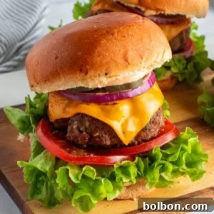 Two cheeseburgers on a wooden board with lettuce, tomato, cheddar cheese, red onion, and pickles, on a brioche bun. A striped napkin is in the background.