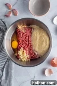 A bowl containing raw ground meat, an egg, minced garlic, and various spices, ready to be mixed. Surrounding the bowl are a cracked eggshell, additional garlic cloves, and an empty mixing bowl. A striped cloth is partially visible on the marble countertop.