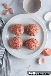 Four raw hamburger patties neatly arranged on a white plate, placed on a marble surface. Nearby, there are fresh garlic cloves, an empty gray bowl, a striped towel, and remnants of cracked eggshells, indicating the preparation stage of burger making.