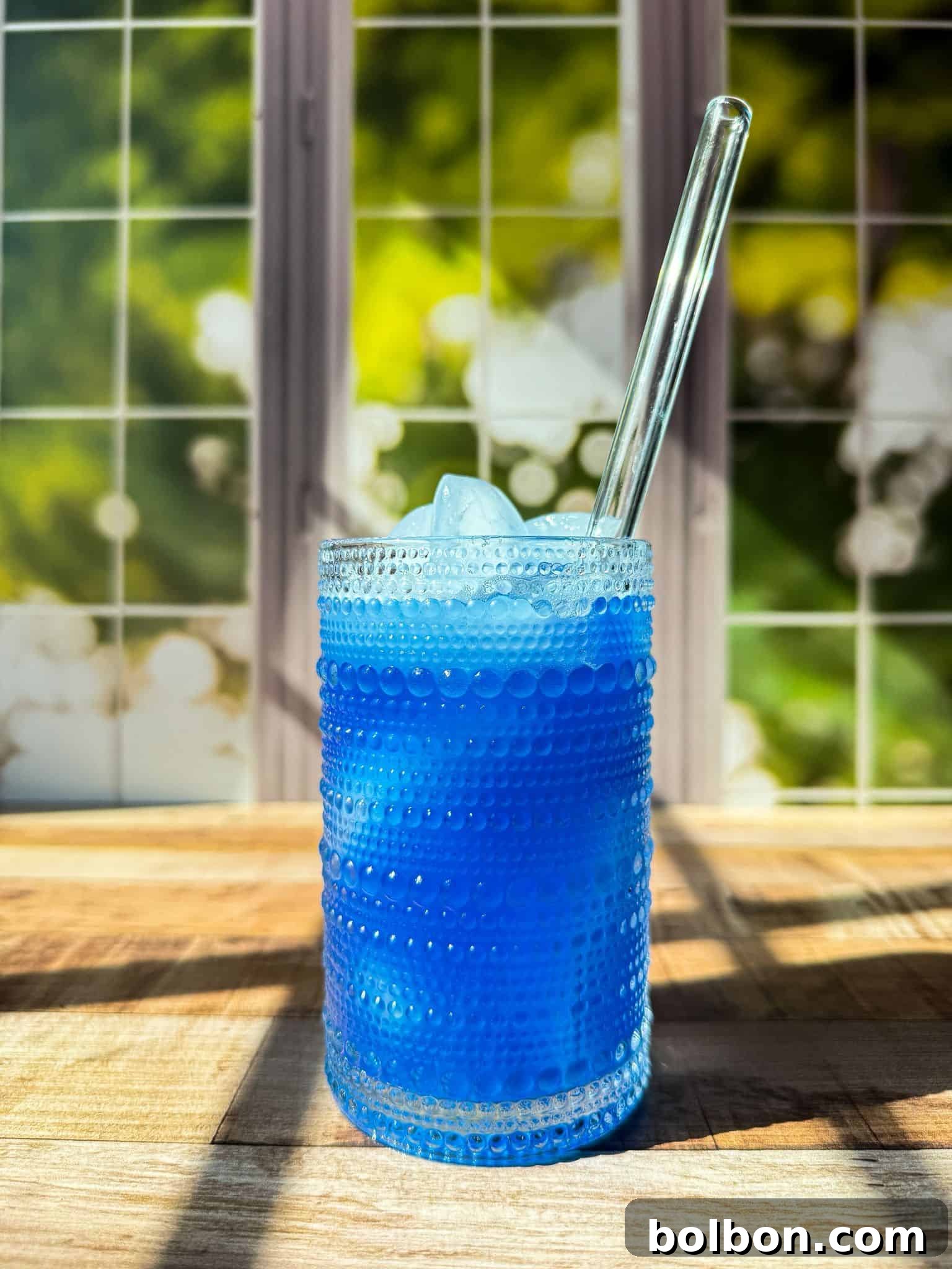 A textured glass filled with homemade gatorade and ice sits on a wooden surface, with a clear straw. Sunlight streams through a window with a green and white background, casting shadows across the scene.