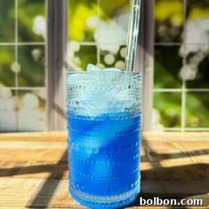 A textured glass filled with blue liquid and ice sits on a wooden surface in sunlight, with a clear straw and a blurred window showing green foliage in the background.
