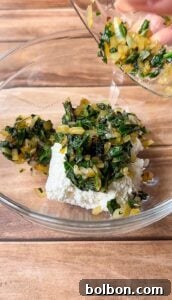 A glass bowl with ricotta cheese topped with sautéed spinach and onions, as more of the mixture is being poured in from another bowl. The scene is set on a wooden surface.