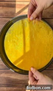 Two hands hold a thin, round sheet of uncooked yellow batter or mixture over a round black tray placed on a wooden surface, with sunlight casting shadows across the scene.