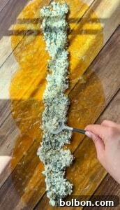 A hand uses a spoon to spread a spinach and cheese filling along the center of overlapping pasta sheets brushed with egg wash on a wooden surface.