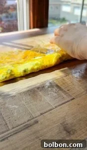 A hand rolls up a thin omelette on a wooden surface by a sunlit window, with some egg spilling onto the board.