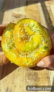 A hand holds a golden, spiral-shaped pastry topped with sesame seeds and green herbs, resting on a wooden surface in sunlight.