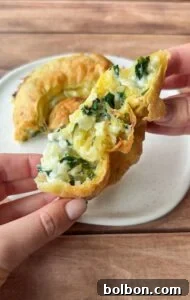A hand holds a flaky pastry filled with creamy cheese and spinach. Another portion of the same pastry sits on a white plate, all on a wooden table background.