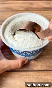 A hand scoops cottage cheese from a container labeled “Good Culture” with a metal 1/2 cup measuring cup on a wooden surface.