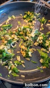 Chopped spinach and onions are being sautéed in a pan, with a sprinkle of salt or seasoning being added from above.