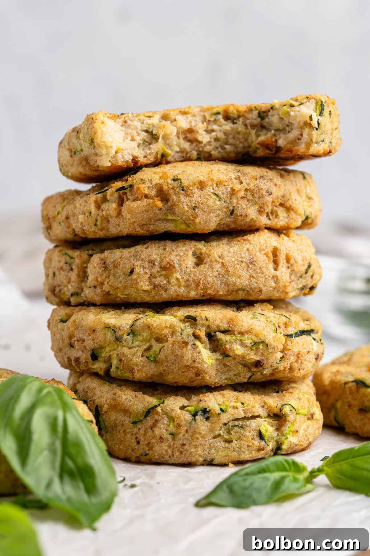 A stack of six zucchini fritters on a white surface, with one fritter on top showing a bite taken out. Fresh basil leaves are scattered around, adding a touch of green to the scene.
