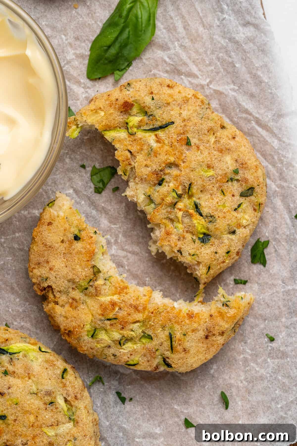 A close-up of a partially eaten zucchini fritter on parchment paper. The fritter is sprinkled with chopped herbs and paired with a small bowl of creamy dip. A basil leaf decorates the scene.