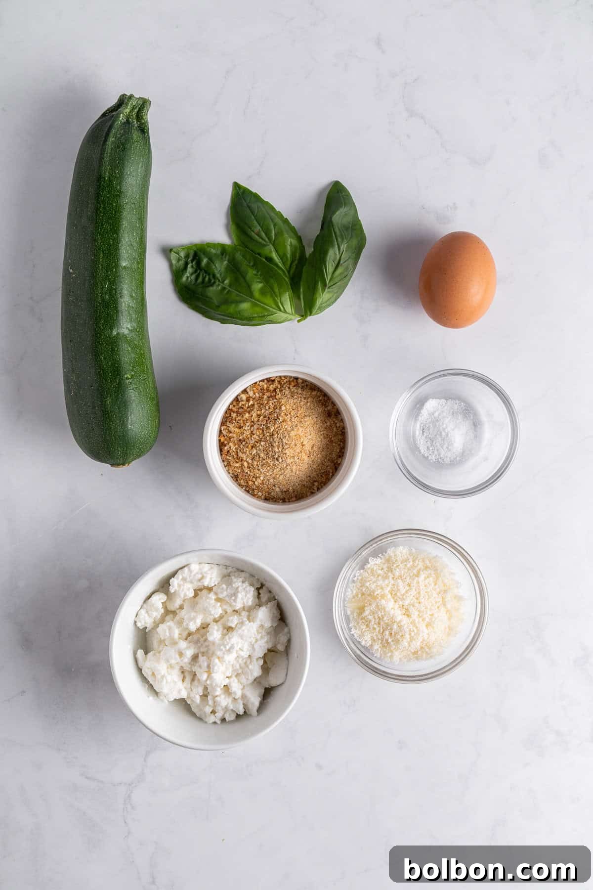 A zucchini, fresh basil leaves, an egg, and small bowls of breadcrumbs, salt, ricotta cheese, and grated Parmesan are arranged on a light marble surface.