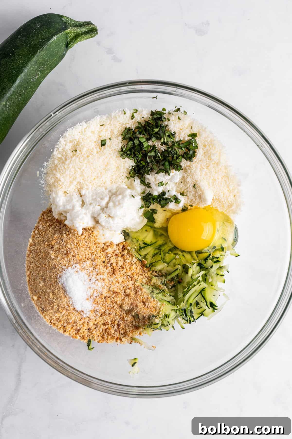A glass bowl containing grated zucchini, an egg, breadcrumbs, grated cheese, chopped herbs, and a bit of white ricotta cheese on a marble surface. A whole zucchini lies beside the bowl.