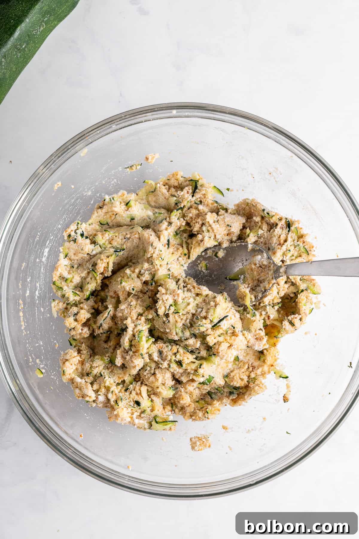 A glass bowl filled with a mixture of grated zucchini and dough, with a metal spoon resting inside. The bowl is placed on a white marble surface, and part of a zucchini is visible in the top left corner.