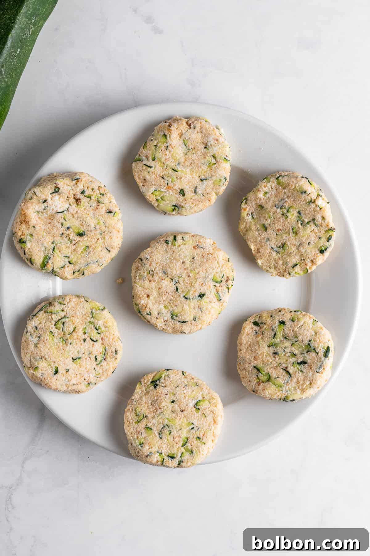 A white plate with seven uncooked, round zucchini fritters, featuring visible zucchini and herbs, placed on a marble countertop.