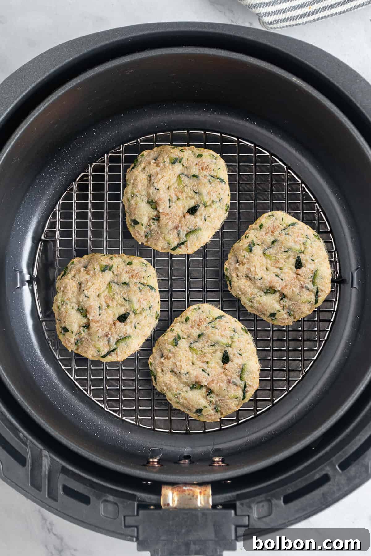 Four zucchini fritters are arranged on a wire rack inside an air fryer basket. They have a golden-brown appearance and are speckled with green zucchini pieces. The background shows part of a gray and white striped cloth.