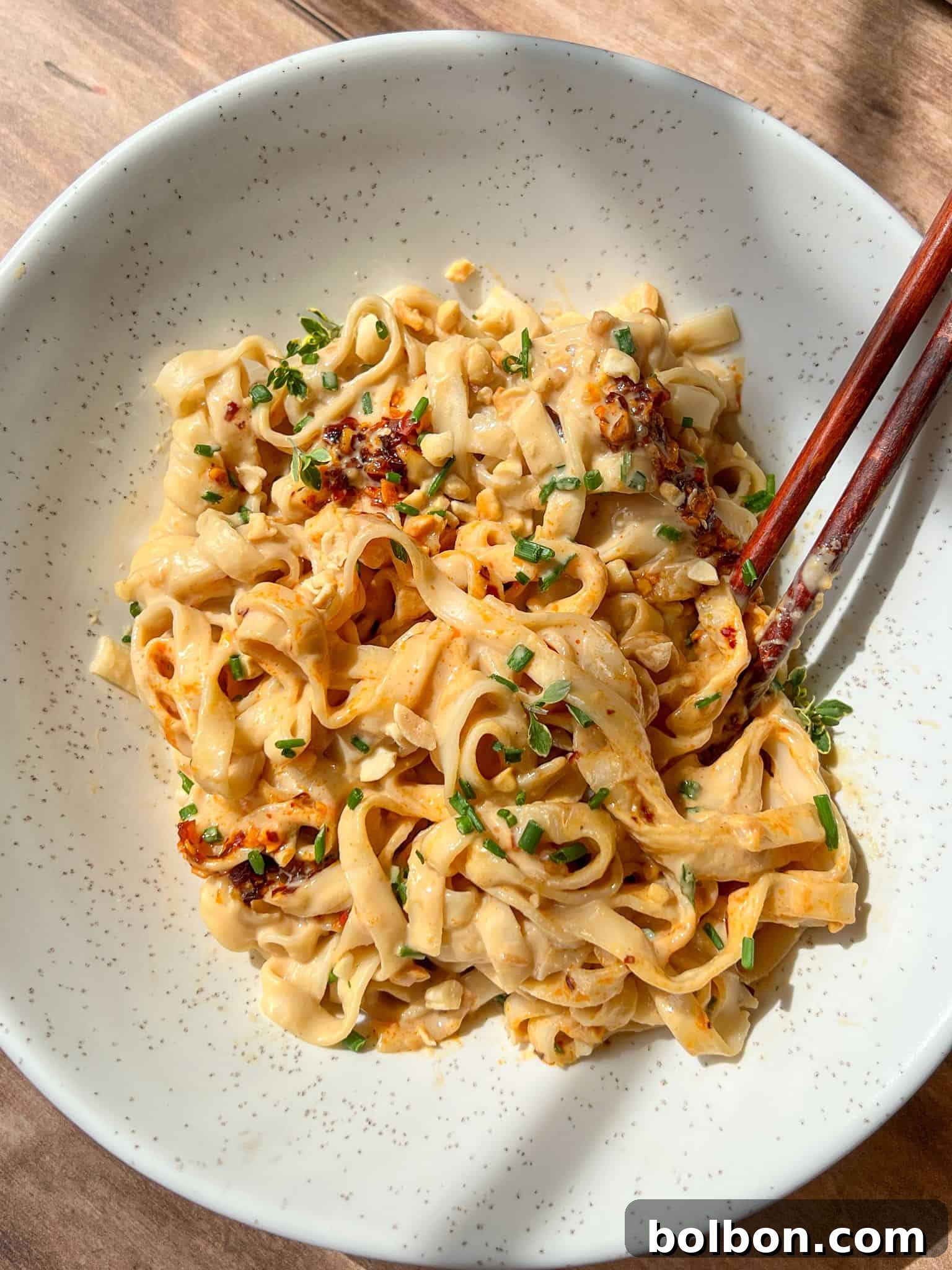 A bowl of wide, flat noodles topped with chili oil, chopped peanuts, and fresh green herbs, with a pair of wooden chopsticks resting on the side of the bowl.
