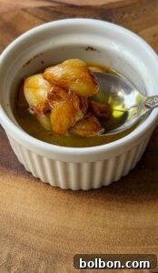 A small white ramekin with roasted garlic cloves in olive oil, resting on a wooden surface. A spoon is placed inside the ramekin, holding some of the caramelized garlic.