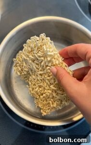 A hand holds a block of uncooked instant ramen noodles over a pot of hot water on a stovetop.