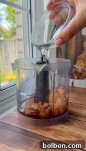 A hand pours clear liquid from a glass bowl into a food processor containing dates, set on a wooden table near a window with sunlight streaming in.