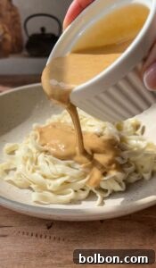 A hand pours creamy brown sauce from a white ramekin onto a plate of cooked fettuccine noodles, set on a wooden surface.
