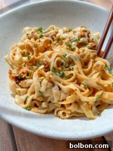 A bowl of wide, flat noodles tossed in a creamy, spicy sauce, garnished with chopped chives. Chopsticks rest on the edge of the bowl. The noodles are served on a light-colored, speckled ceramic dish.