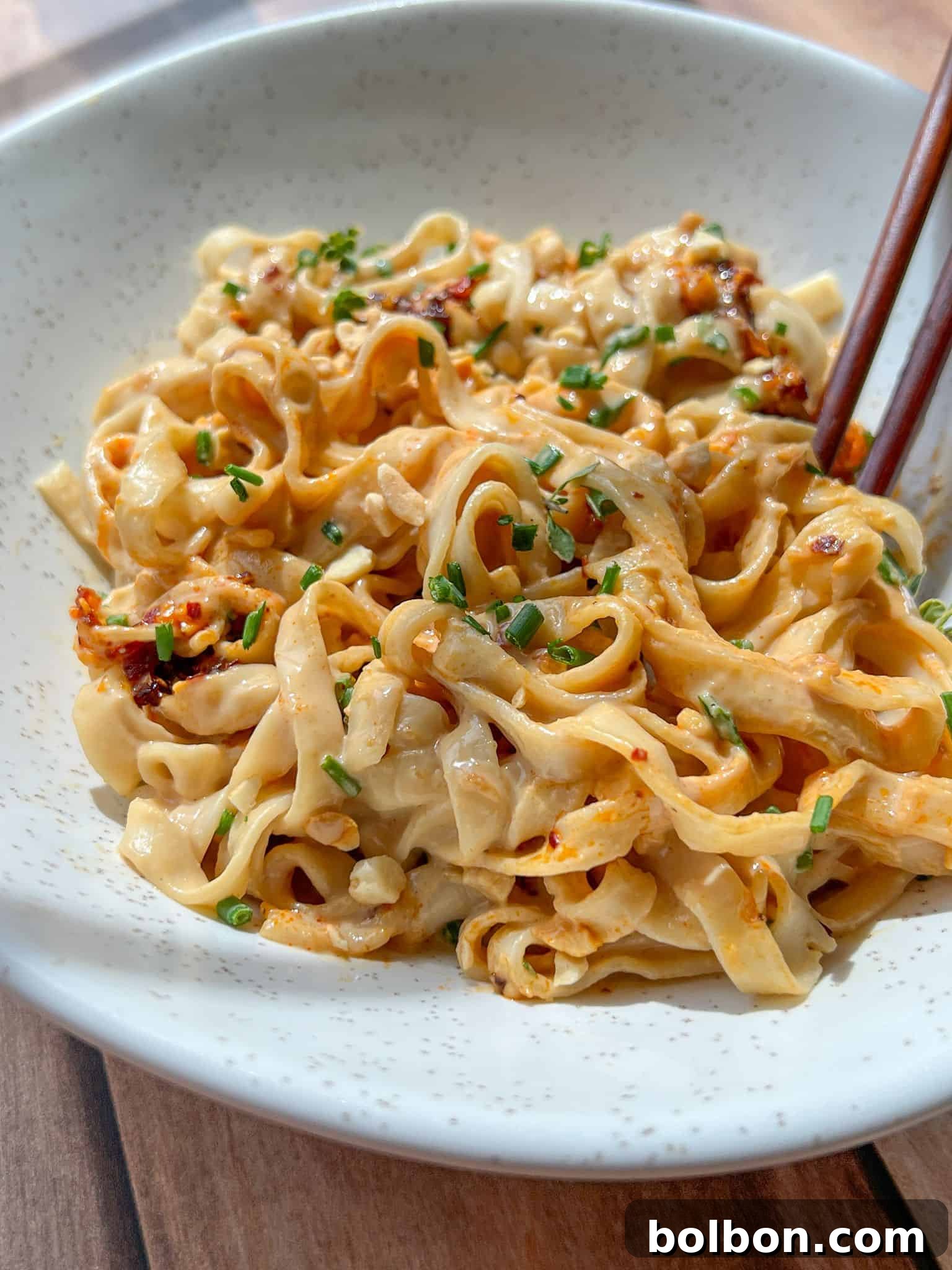 A bowl of wide, flat noodles tossed in a creamy, spicy sauce, garnished with chopped chives. Chopsticks rest on the edge of the bowl. The noodles are served on a light-colored, speckled ceramic dish.