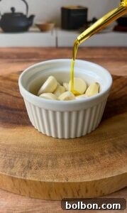 A white ramekin filled with peeled garlic cloves sits on a wooden board, while golden olive oil is being drizzled over the garlic from above with a spoon. Kitchen items are blurred in the background.
