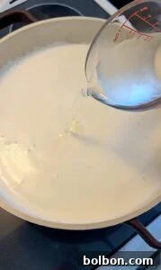 Craft Your Own Creamy Cottage Cheese 6 A clear liquid is being poured from a measuring cup into a large pot filled with milk or cream on a stovetop.