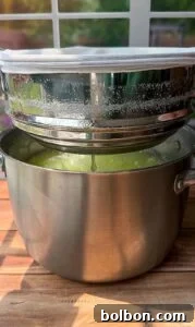 Craft Your Own Creamy Cottage Cheese 8 A metal colander lined with a white cloth sits over a stainless steel pot, with liquid dripping through. Sunlight streams onto the wooden surface beneath.
