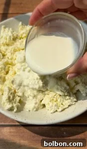 Craft Your Own Creamy Cottage Cheese 10 A hand pours milk from a small glass bowl over a plate of crumbled cheese, set on a wooden table.