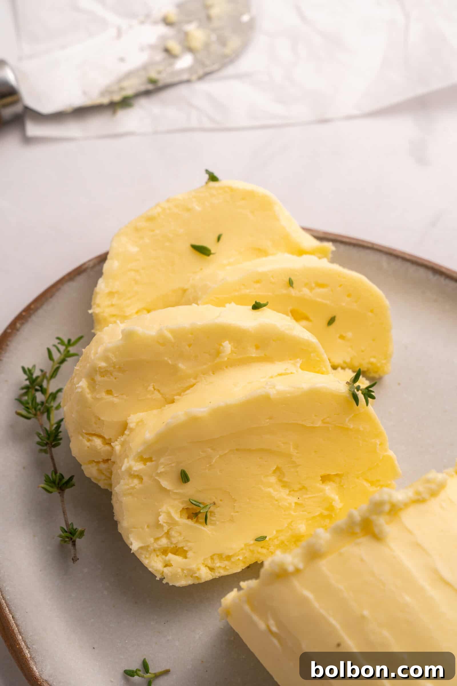 Churning Your Own Golden Butter 2 Sliced homemade butter with sprigs of fresh thyme on a ceramic plate, with a butter knife and parchment paper in the background.