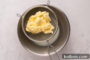 Churning Your Own Golden Butter 7 A metal strainer holding fresh yellow butter sits over a stainless steel bowl containing liquid buttermilk on a light marble surface.