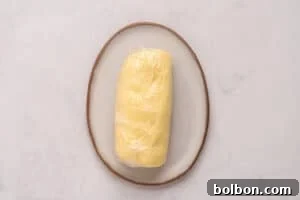 Churning Your Own Golden Butter 9 A log of yellow dough wrapped in plastic wrap sits on an oval ceramic plate with a brown rim, placed on a light marble surface.