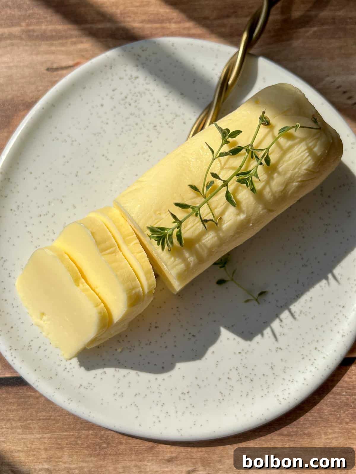 Churning Your Own Golden Butter 3 A log of sliced butter garnished with fresh thyme sits on a speckled white plate, with a gold-handled knife beside it, all on a wooden surface lit by sunlight.