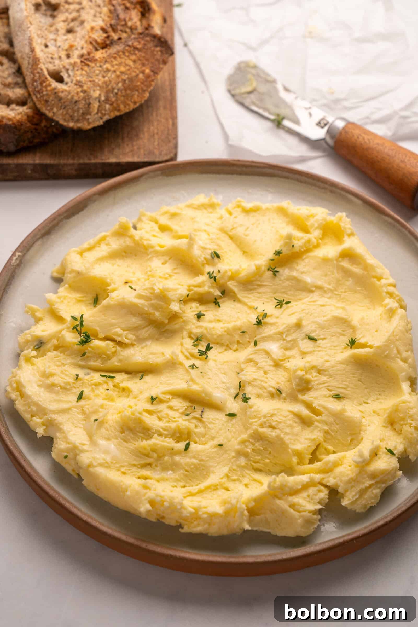 Churning Your Own Golden Butter 4 A plate of creamy, whipped butter garnished with fresh herbs sits on a table next to slices of rustic bread and a butter knife.