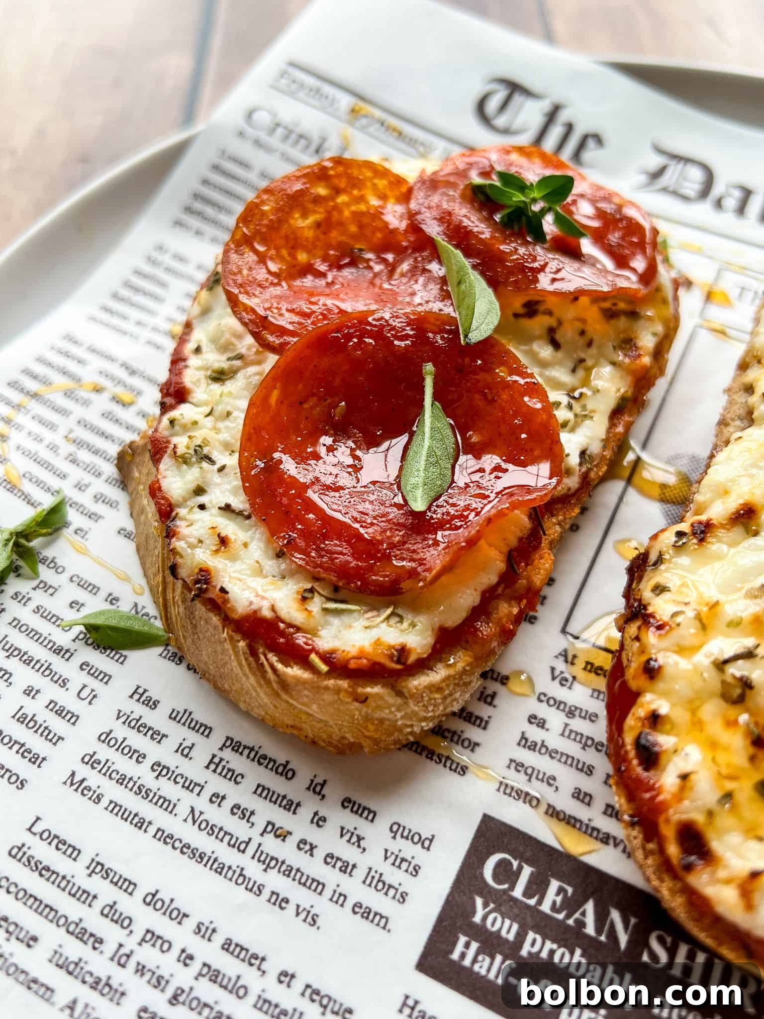 Creamy Cottage Cheese Toast Pizzas 2 A slice of bread topped with melted cheese, pepperoni slices, and fresh herbs sits on a plate lined with newspaper-style parchment paper. Another similar slice is partially visible beside it.