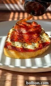 A slice of bread topped with cheese, tomato sauce, and pepperoni is served on a plate while honey is being drizzled over it. Sunlight and shadows create a striped pattern across the scene.
