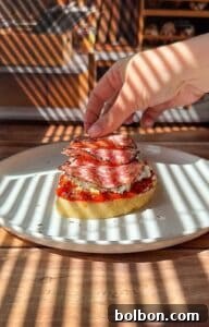 A hand places slices of salami on top of bread with red sauce and cheese, on a white plate. Sunlight streams through blinds, casting striped shadows over the food and table.