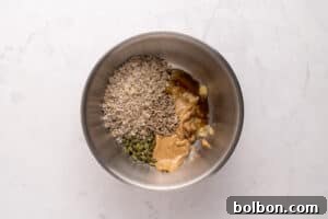 A metal bowl containing rolled oats, sunflower seeds, pumpkin seeds, peanut butter, honey, and mashed banana on a light marble surface.