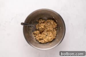 A metal mixing bowl with a spoon inside, containing a thick, mixed batter or dough with visible grains and herbs, placed on a light marble surface.