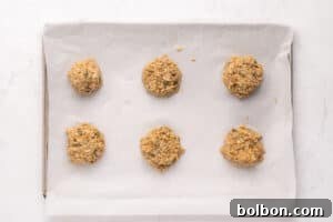 Six scoops of unbaked cookie dough spaced evenly on a parchment-lined baking sheet, ready to be baked, set on a white marble surface.
