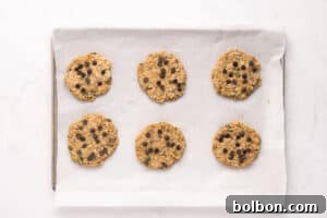 Six unbaked chocolate chip cookies are arranged on a baking sheet lined with parchment paper, ready to be baked. The cookies are spaced evenly apart on a white surface.
